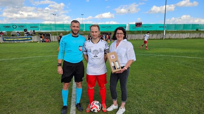 Drei Personen stehen auf einem Fußballfeld mit einem Fußball. Ein Mann in roten Shorts hält eine Trophäe.