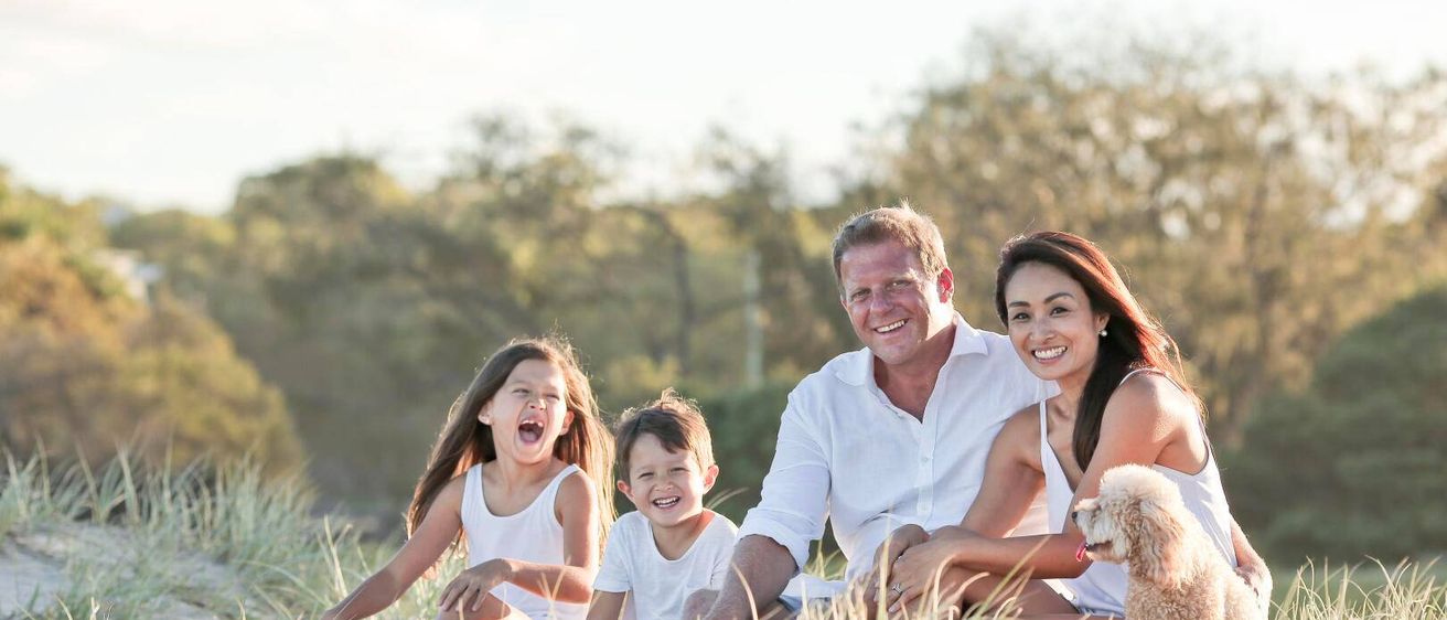 Eine vierköpfige Familie lächelt und sitzt auf einem Grasfeld. Die Frau trägt ein ärmelloses Kleid und der Mann ein weißes Hemd.