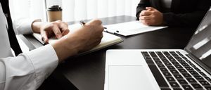 Zwei Personen in einer Besprechung, eine schreibt auf ein Notizbuch, ein Laptop und eine Tasse stehen auf dem Tisch.