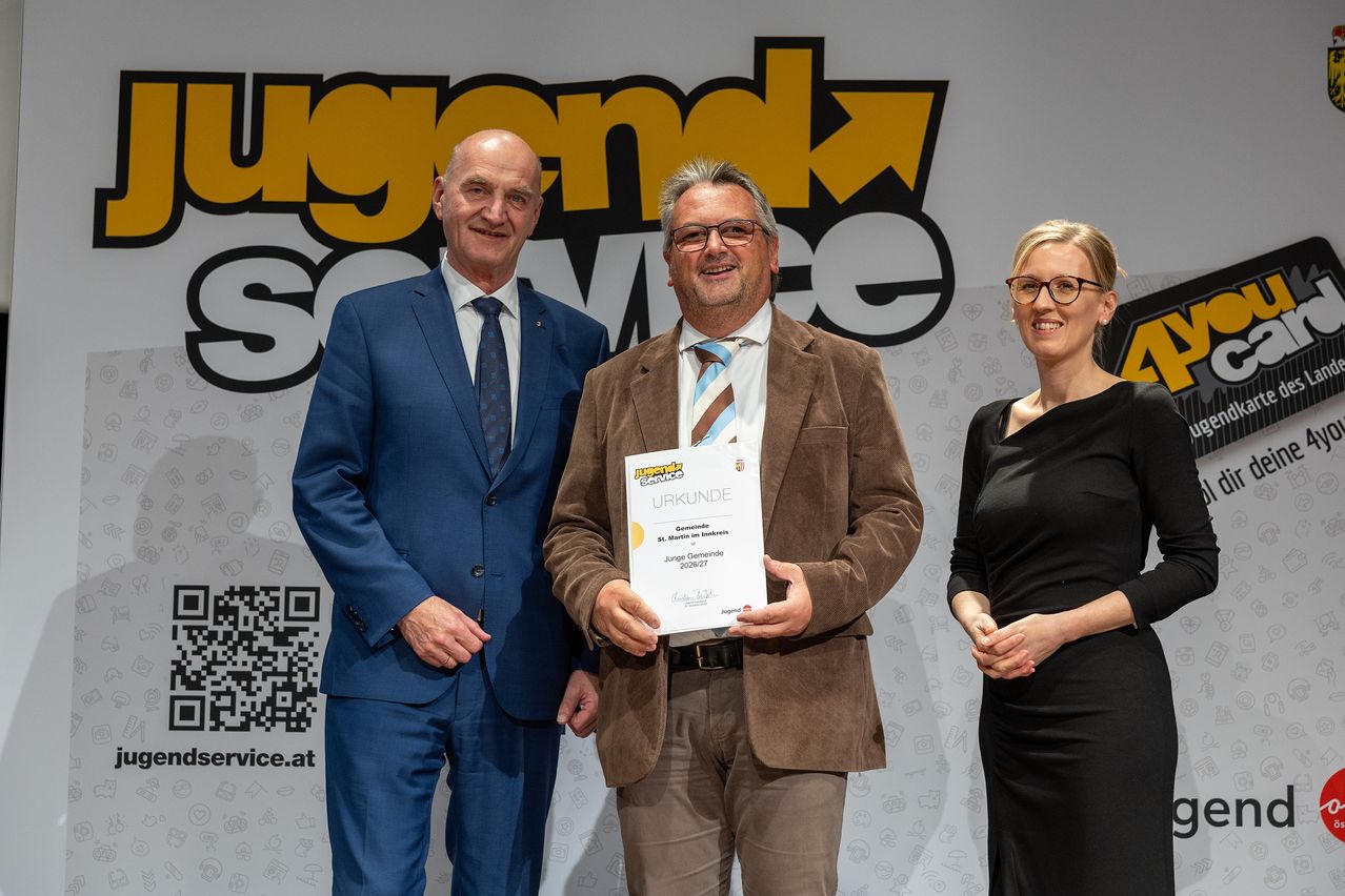 Drei Personen stehen zusammen vor einer Wand mit einem Schild, auf dem 'jugendk' und 'swe' steht. Der Mann in der Mitte hält ein Zertifikat und trägt einen braunen Anzug und eine Brille. Der Mann auf der linken Seite trägt einen blauen Anzug und eine Krawatte. Die Frau auf der rechten Seite trägt ein schwarzes Kleid und eine Brille.