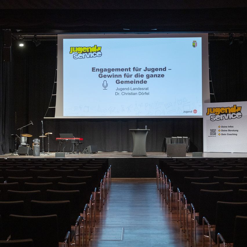 Ein Auditorium mit einer Bühne, auf der eine Präsentation mit dem Titel 'Engagement für Jugend - Gewinn für die ganze Gemeinde' von Jugend-Landesrat Dr. Christian Dorfel angezeigt wird. Musikinstrumente und Lautsprecher sind auf der Bühne aufgebaut.