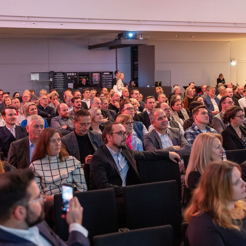 Ein großes Publikum in formeller Kleidung sitzt in Reihen, wahrscheinlich bei einer Konferenz. Ein Mann in der vorderen Reihe macht ein Foto mit seinem Handy.