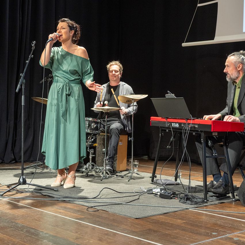 Eine Frau in einem grünen Kleid singt in ein Mikrofon auf der Bühne, begleitet von einem Mann am Keyboard und einem anderen am Schlagzeug.