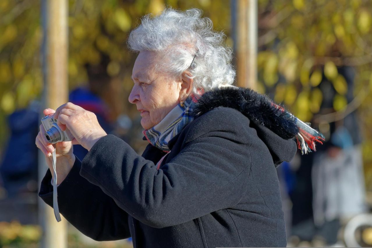 Eine ältere Frau in einem schwarzen Mantel mit einer mit Fell gefütterten Kapuze macht ein Foto mit ihrer Kamera im Freien.