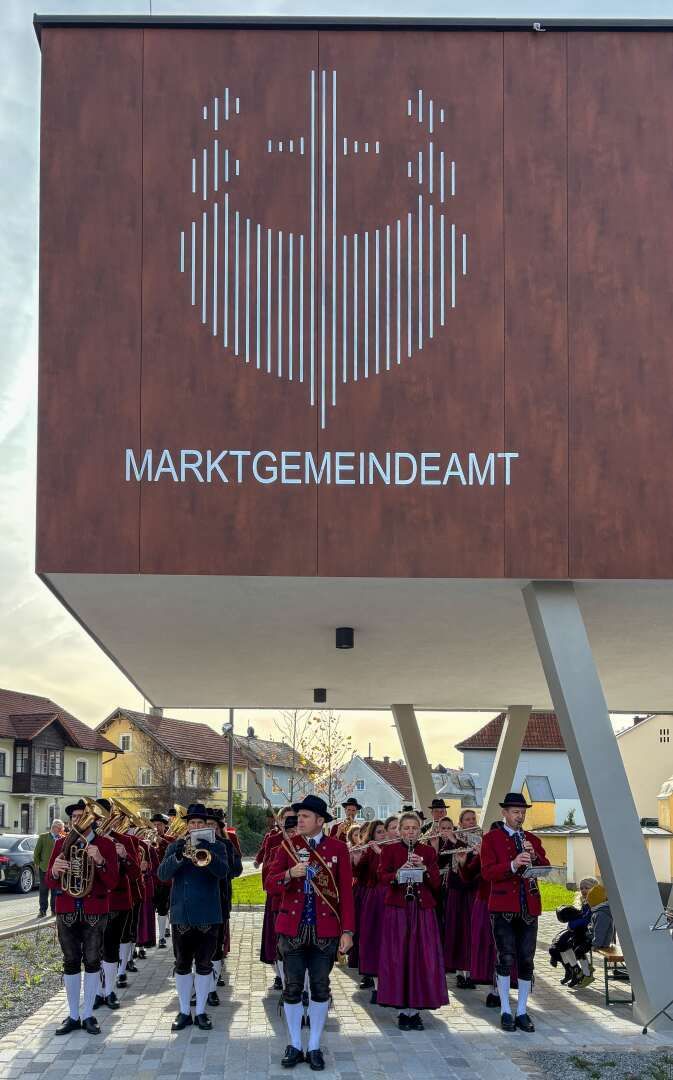 Eine Gruppe von Musikern in roten Uniformen und Hüten spielt Instrumente vor einem Gebäude mit einer braunen Wand und weißem Text. Das Bild zeigt die Fassade des Gebäudes und ein großes Logo.