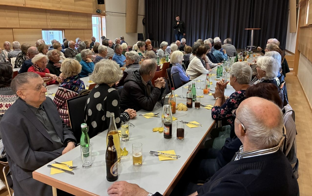 Ein Saal mit Bühne, Redner und Publikum, das an Tischen sitzt und eine Aufführung beobachtet. Flaschen und Gläser stehen auf den Tischen. Das Publikum ist älter.
