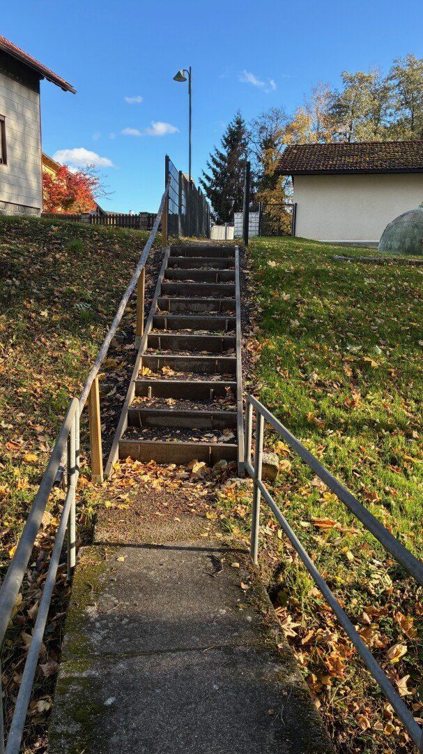 Betonierte Stufen mit Metallgeländern führen einen Grashügel hinauf. Im Hintergrund befindet sich ein Haus mit einem Ziegeldach und einem kleinen Baum.