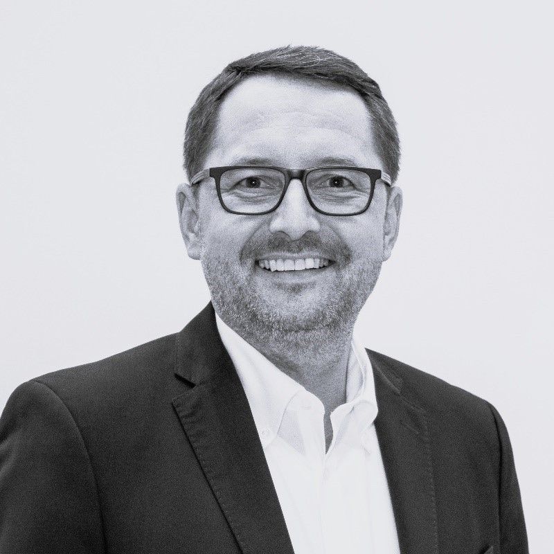 Ein Mann mit Bart und Brille, gekleidet in einen schwarzen Anzug und weißes Hemd, lächelt direkt in die Kamera.