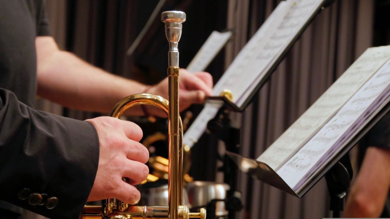 Ein Musiker spielt eine Trompete, hält sie mit einer Hand und liest mit der anderen Hand Noten. Die Trompete ist golden und die Noten sind weiß.
