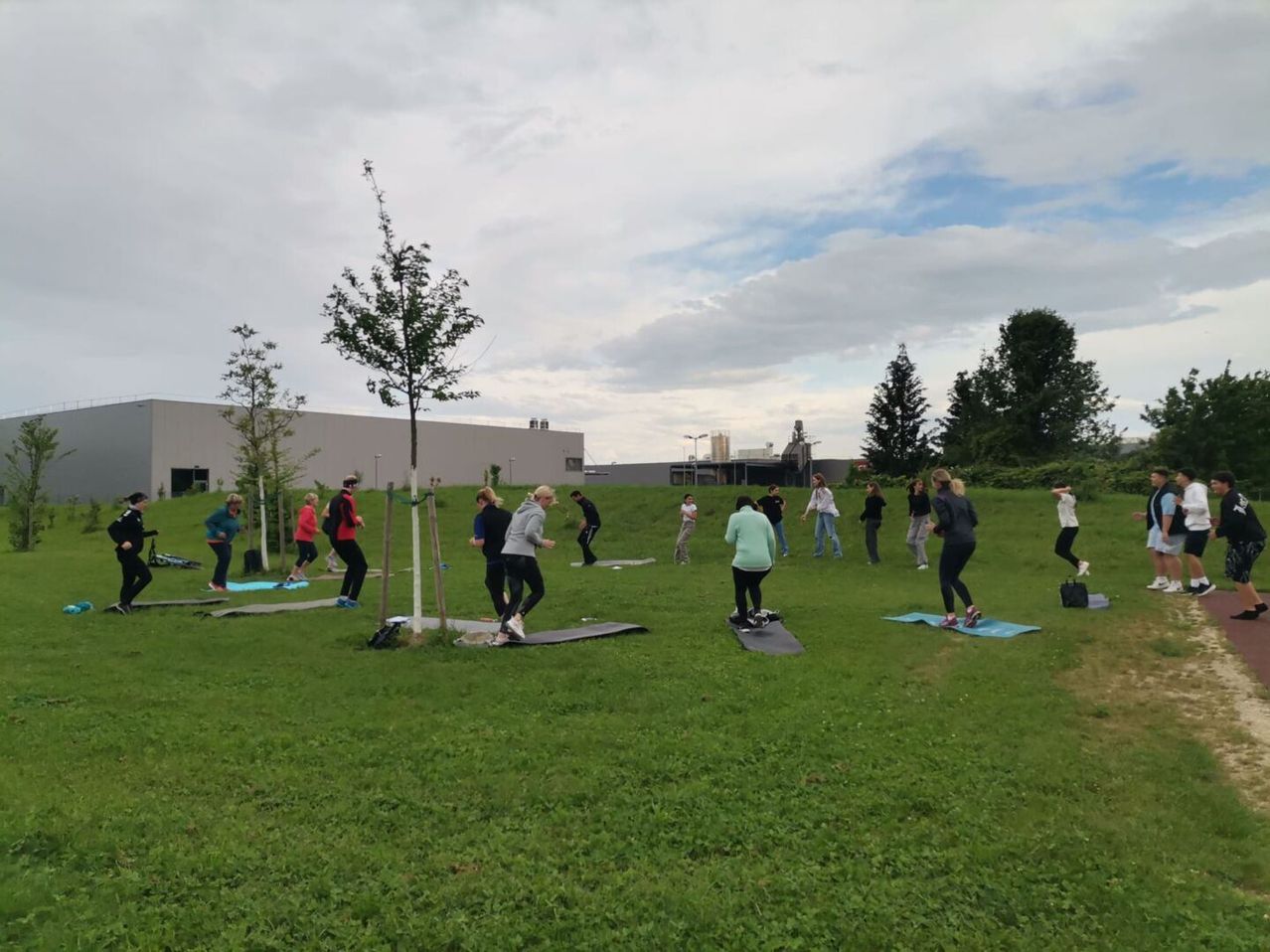 Eine Gruppe von Menschen praktiziert Yoga auf einem Rasenfeld, einige stehend und einige auf Yogamatten sitzend. Bäume und Büsche sind in der Nähe, und ein Gebäude ist in der Ferne zu sehen.