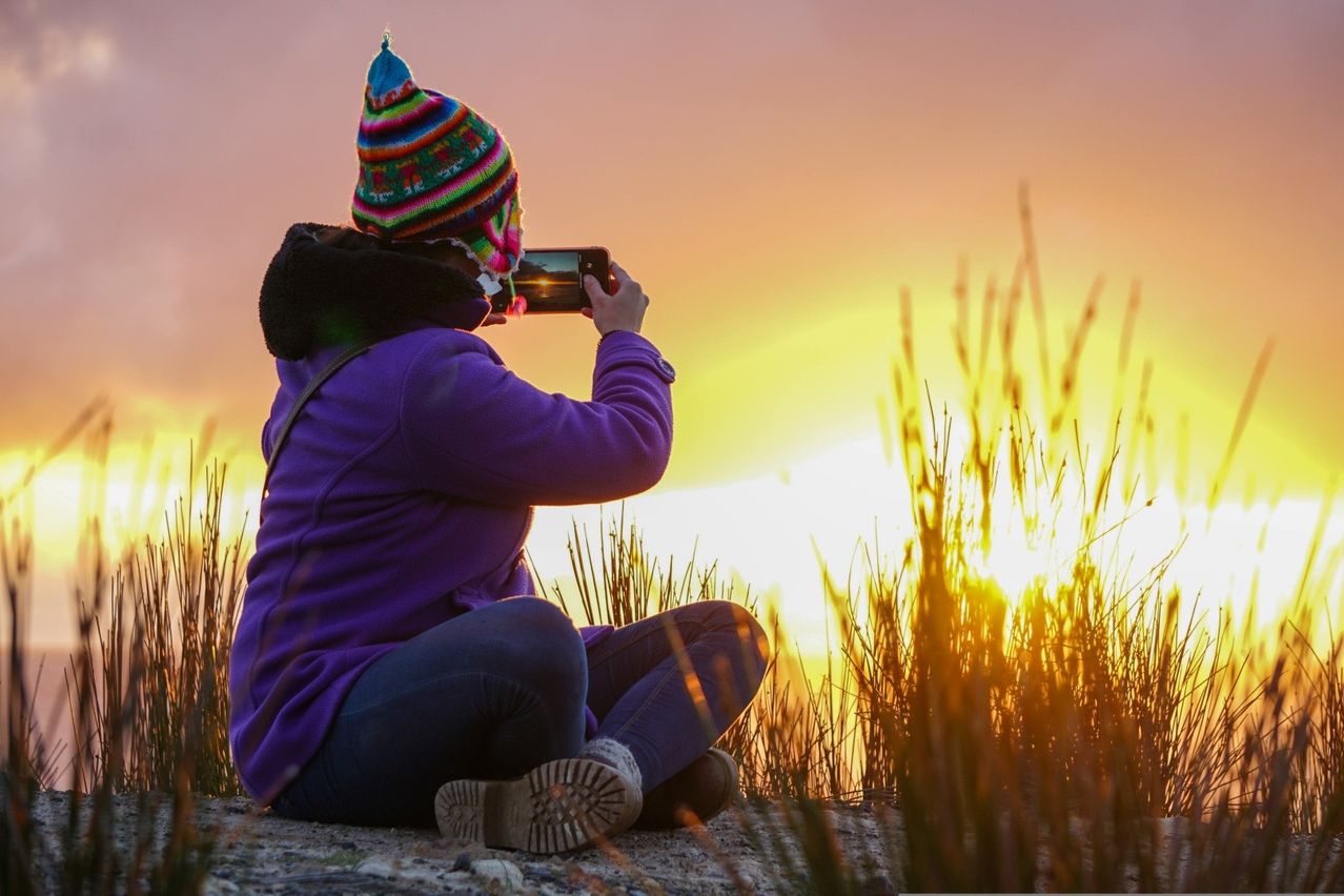Eine Frau sitzt auf dem Boden und fotografiert einen Sonnenuntergang mit ihrem Handy. Sie trägt einen bunten Hut und eine lila Jacke.