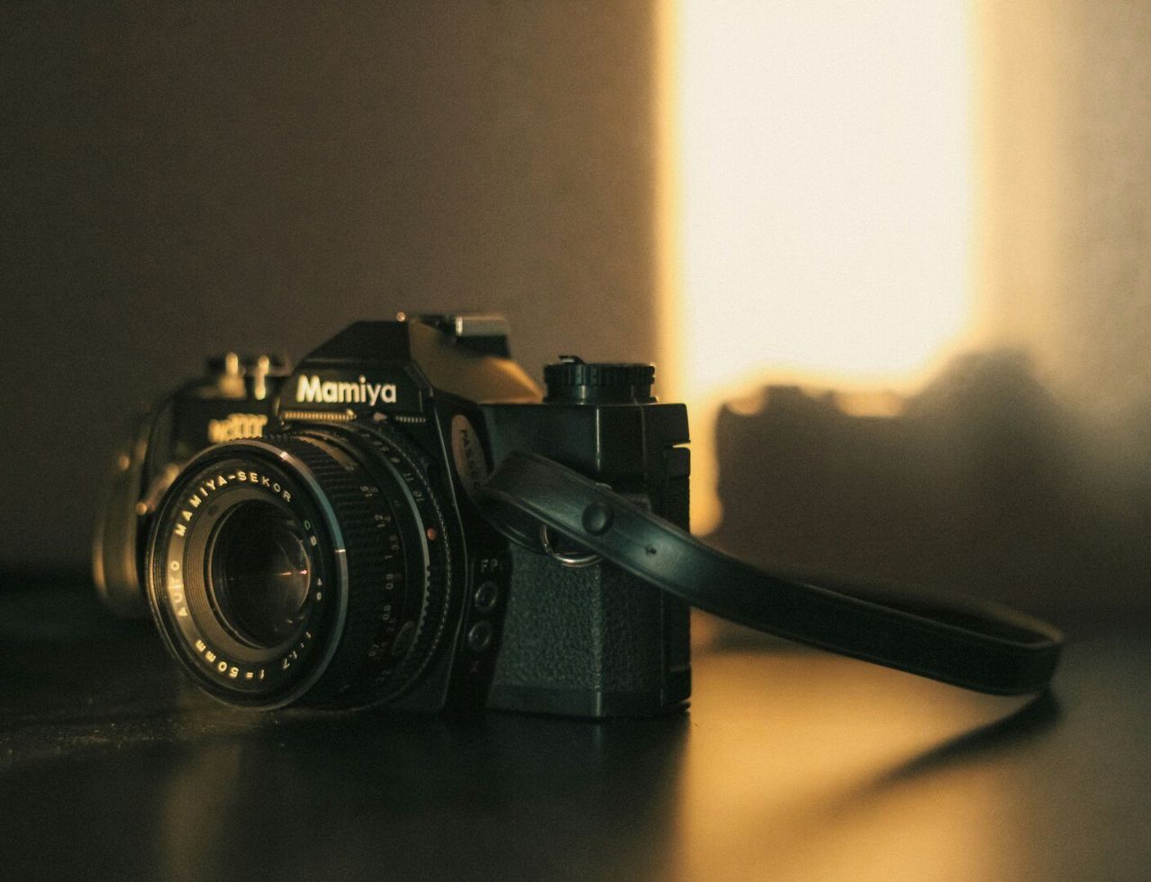 Eine schwarze Mamiya-Kamera mit einem Riemen liegt auf einer dunklen Oberfläche, möglicherweise einem Tisch, mit Licht, das von der rechten Seite scheint.