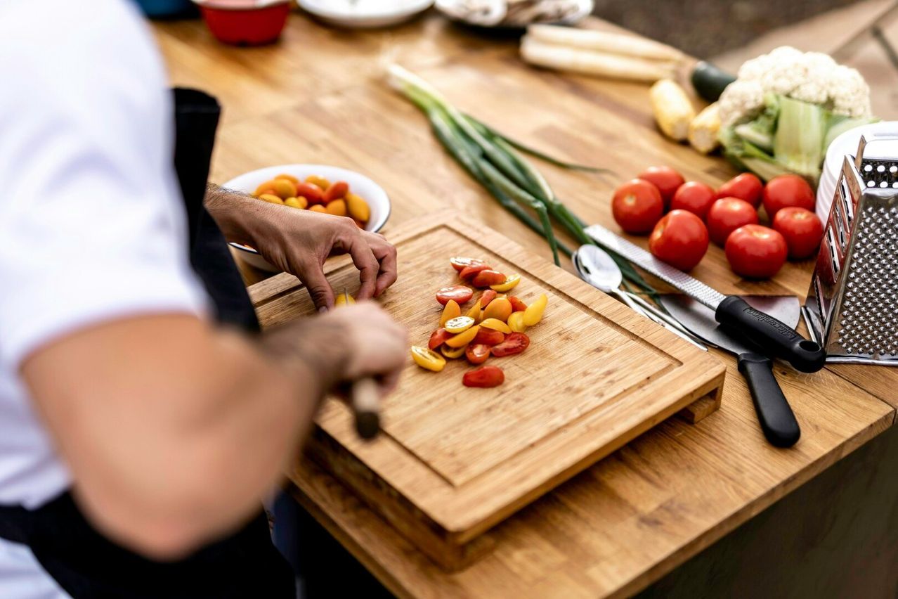 Ein Koch bereitet Tomaten auf einem Bambusschneidebrett auf einem Holztisch mit verschiedenen Gemüsesorten zu.