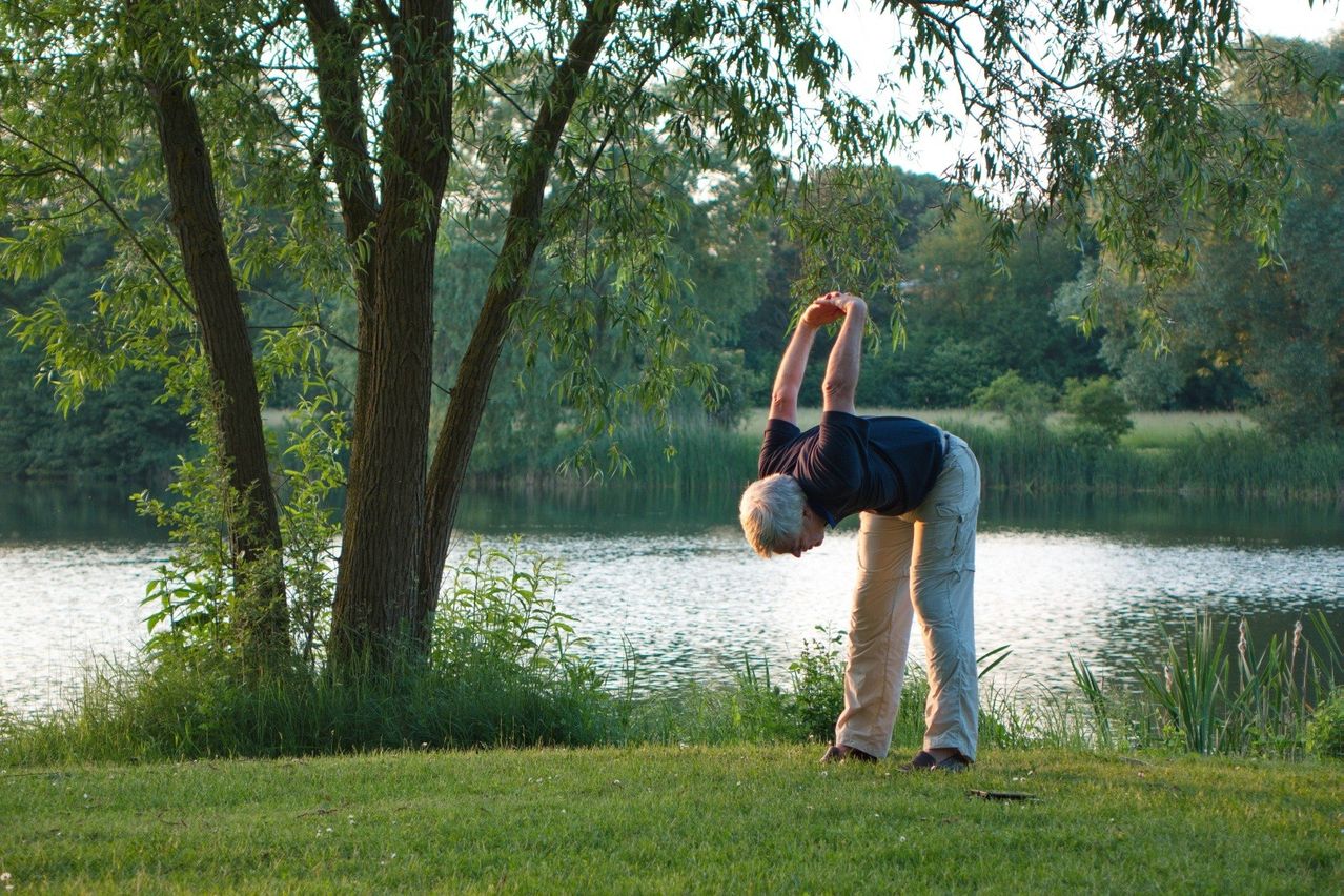 Ein älterer Mann macht eine Yoga-Pose in der Nähe eines Sees, mit einem Baum und Grünpflanzen im Hintergrund.