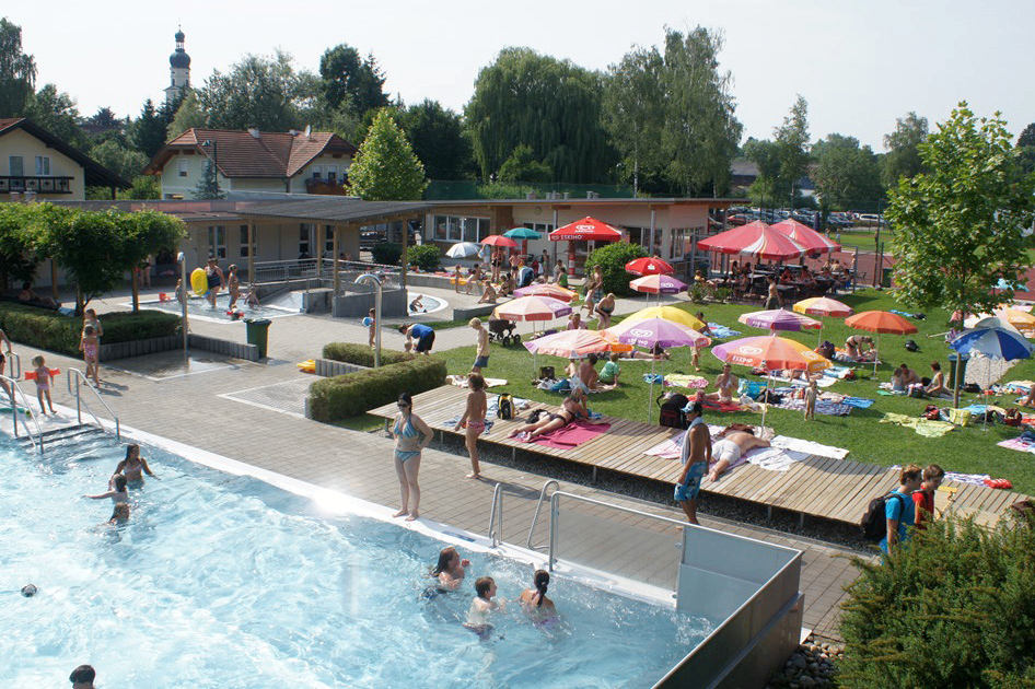 Ein großer Außenpoolbereich mit vielen Menschen, die schwimmen und sich unter bunten Sonnenschirmen entspannen. Gebäude und Bäume im Hintergrund.