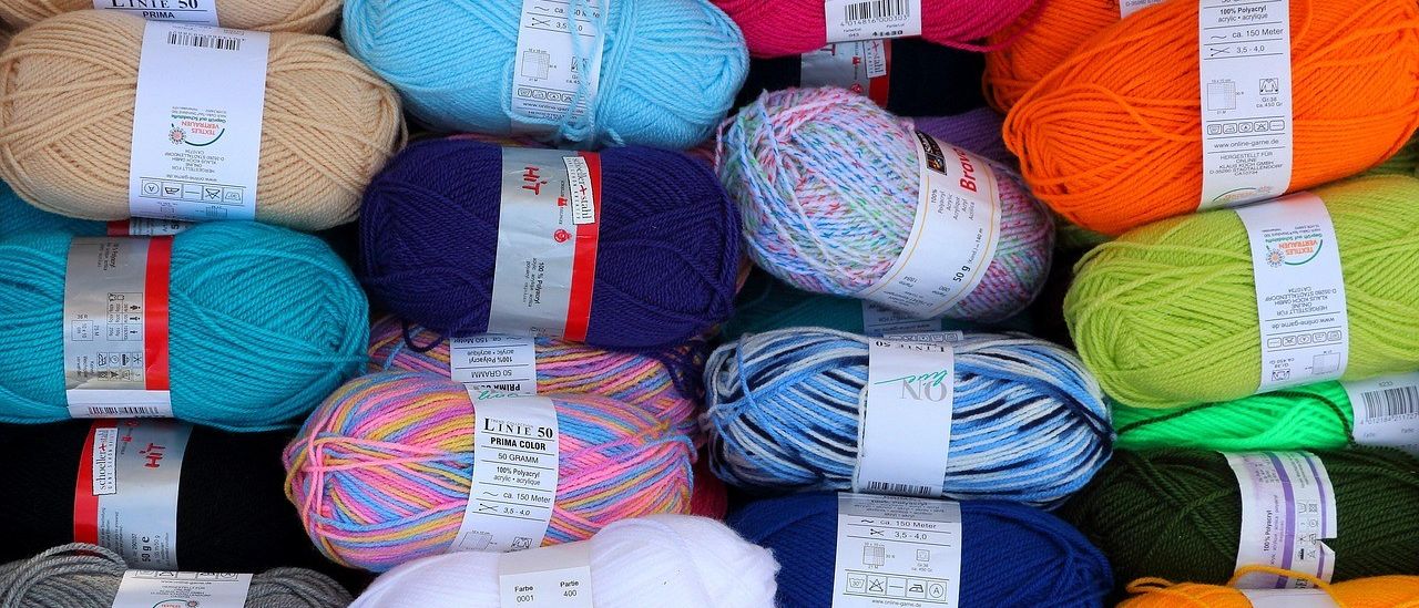 Verschiedene Garnrollen in mehreren Farben und Schattierungen sind zusammengestapelt. Jede Kugel hat ein Etikett mit Produktdetails wie Gewicht, Material und Farbname.