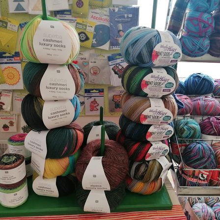 Eine Anzeige von verschiedenen Garnkugeln in verschiedenen Farben und Marken, wie Superba und Woolly Hugs, sind in einem Geschäft mit einem gelb-rot gestreiften Boden gestapelt und organisiert.