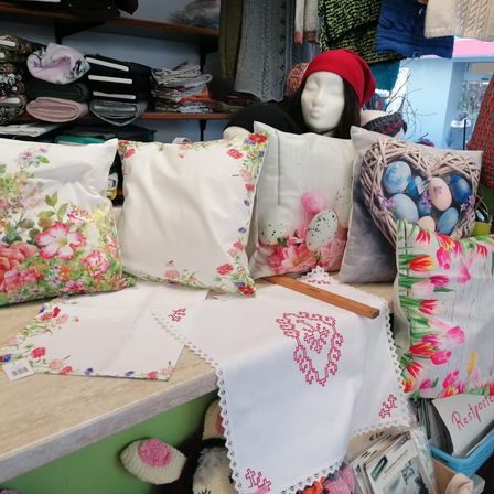 Ein Geschäft zeigt dekorative Kissen mit Blumen- und Ostereier-Designs auf einem Tresen. Ein Mannequin-Kopf mit einer roten Mütze steht hinter dem Tresen. Verschiedene Stoffe hängen an der Wand und ein Korb hält ein 'Restposte'-Schild.