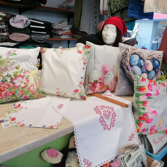 Ein Geschäft zeigt dekorative Kissen mit Blumen- und Ostereier-Designs auf einem Tresen. Ein Mannequin-Kopf mit einer roten Mütze steht hinter dem Tresen. Verschiedene Stoffe hängen an der Wand und ein Korb hält ein 'Restposte'-Schild.