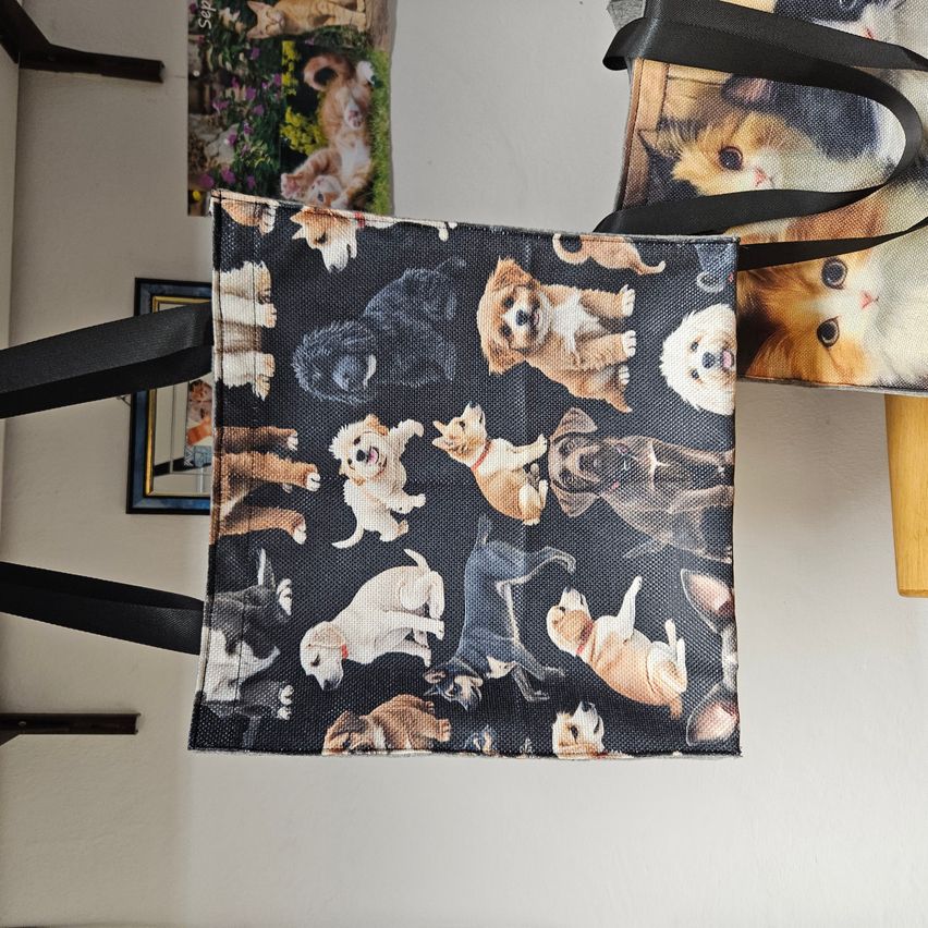 Ein Stoffbeutel mit verschiedenen Hunde- und Katzendesigns, der an schwarzen Henkeln hängt. Dahinter ist teilweise ein weiterer Beutel mit Katzendesigns zu sehen.