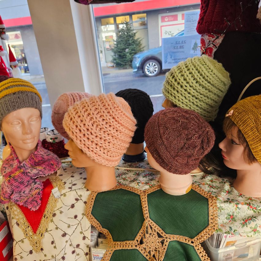 Manequins mit verschiedenen gestrickten Mützen und Schals in einem Schaufenster ausgestellt. Verschiedene Farben der Mützen sind sichtbar, darunter Gelb, Rosa, Grün und Braun.