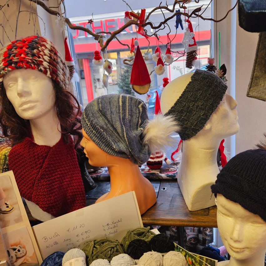 Ein Tisch zeigt verschiedene gestrickte Mützen und Beanies auf Mannequinköpfen. Die Mützen umfassen eine rote Mütze, eine grau-weiß gestreifte Mütze und eine schwarze Mütze. Ein Schild mit Preisen und ein Haufen Wolle sind ebenfalls zu sehen.