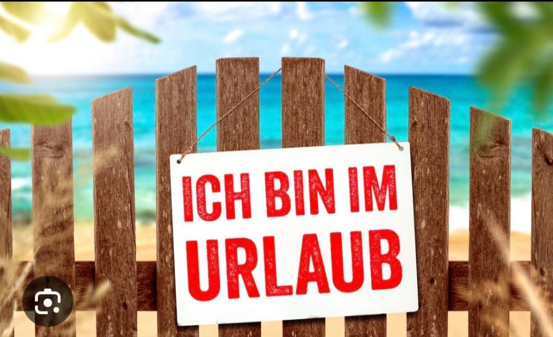 Ein weißes Schild mit rotem Text, das 'ICH BIN IM URLAUB' steht, hängt an einem Holzzaun mit Strand und Meer im Hintergrund.