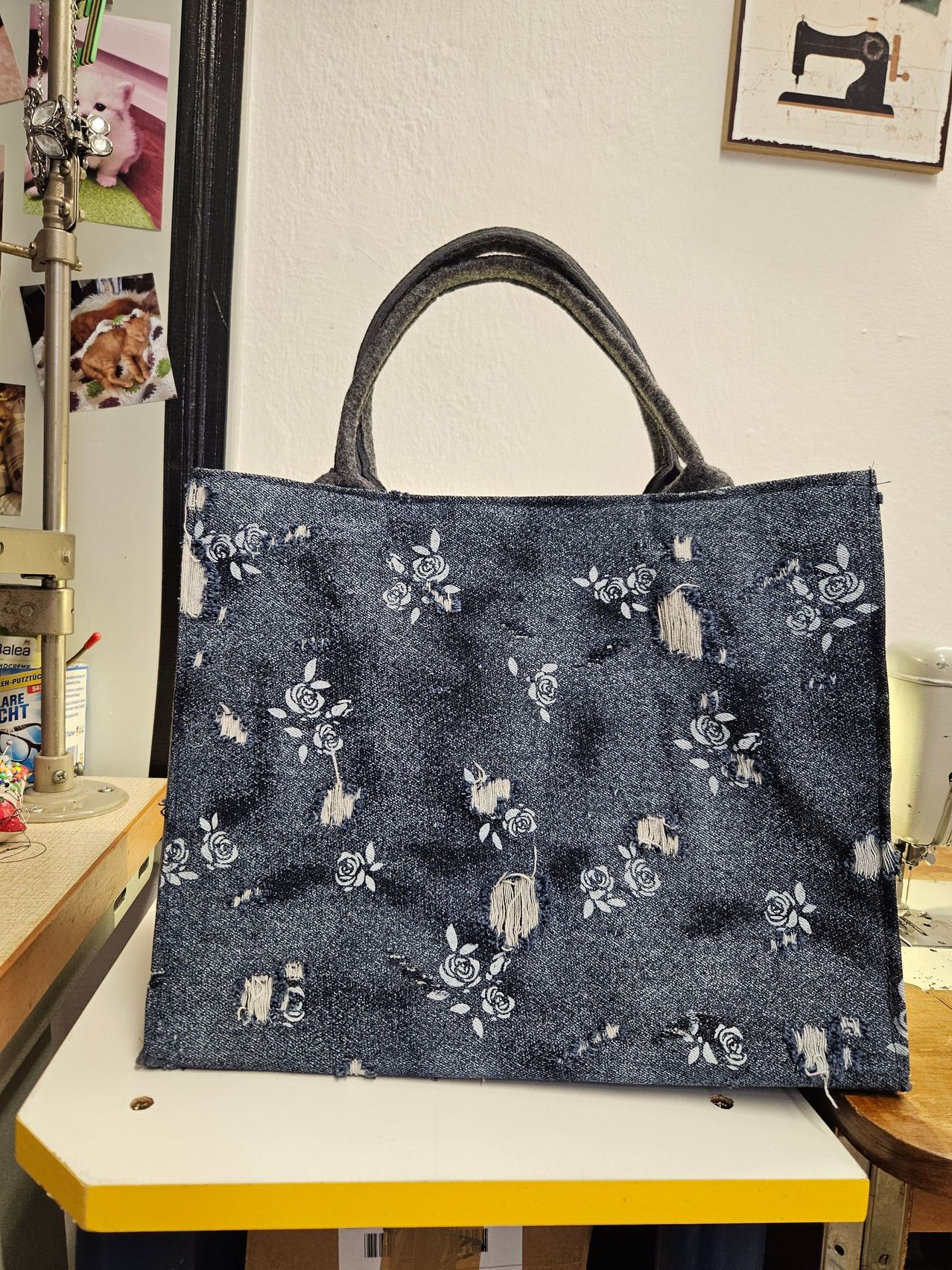 A denim tote bag with a distressed floral pattern on a white background, placed on a table with a white wall in the background.