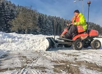 Ein Mann in einer gelben und orangefarbenen Jacke benutzt einen an einen roten Rasenmäher angehängten Schneefräse, um Schnee von der Straße zu räumen.