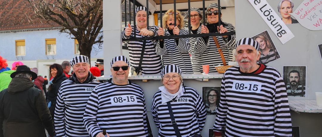 Eine Gruppe von Menschen in schwarz-weiß gestreiften Gefängnisuniformen posiert für ein Foto in einer nachgestellten Gefängniszelle. Einige haben falsche Schnurrbärte. Der Mann in der Mitte hält eine Bierflasche.