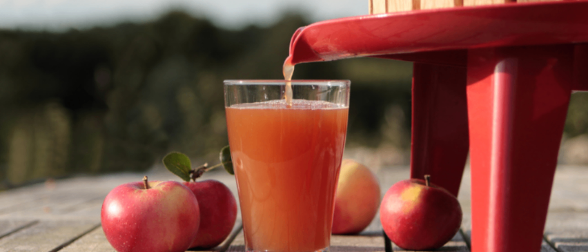 Ein Glas frisch gepresster Apfelsaft wird aus einer roten Apfelpresse gegossen, umringt von Äpfeln.