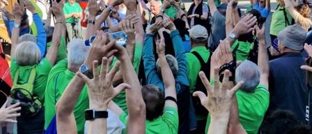 Eine Menschenmenge in grünen Hemden und Mützen hebt ihre Hände bei einer Versammlung, wahrscheinlich an einer Protestaktion teilnehmend. Im Hintergrund sind ein rotes Fahrzeug und einige Autos geparkt.