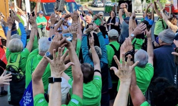 Eine Menschenmenge in grünen T-Shirts hebt ihre Hände in die Luft, vermutlich bei einer Veranstaltung im Freien. Einige tragen Hüte und Uhren, und eine Frau in der Mitte hat ihr Haar zu einem Zopf gebunden. Fahrzeuge und Personen sind im Hintergrund zu sehen.