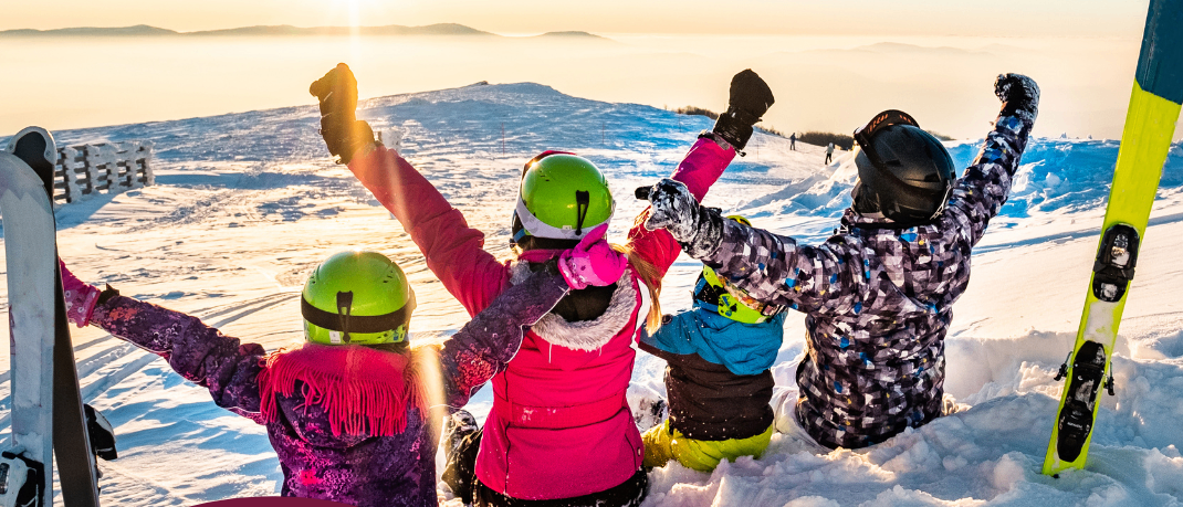Vier Kinder sitzen mit ihren Skiern und Helmen im Schnee. Sie heben ihre Hände. Das Bild hat einen Wasserzeichen von 17.01. & 19.02.2026. Der Hintergrund ist ein Sonnenuntergang.