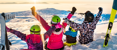 Vier Kinder sitzen mit ihren Skiern und Helmen im Schnee. Sie heben ihre Hände. Das Bild hat einen Wasserzeichen von 17.01. & 19.02.2026. Der Hintergrund ist ein Sonnenuntergang.