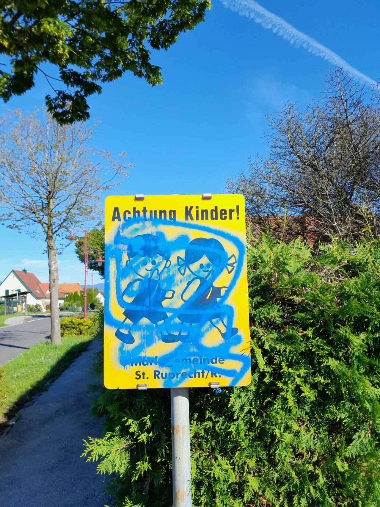 Ein gelbes Schild mit blauer Graffiti warnt vor Kindern. Es steht an einem Pfosten neben einer Hecke, mit einem Haus und Bäumen im Hintergrund.