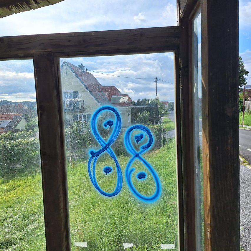 Eine blaue Zeichnung eines Unendlichkeitssymbols ist auf einem Glasfenster. Hinter dem Fenster befindet sich ein Grasfeld und Häuser.