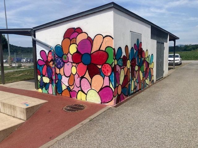 Ein farbenfrohes Blumen-Wandbild schmückt die Wand eines kleinen Gebäudes in der Nähe eines Parkplatzes. Ein weißes Auto ist rechts geparkt.