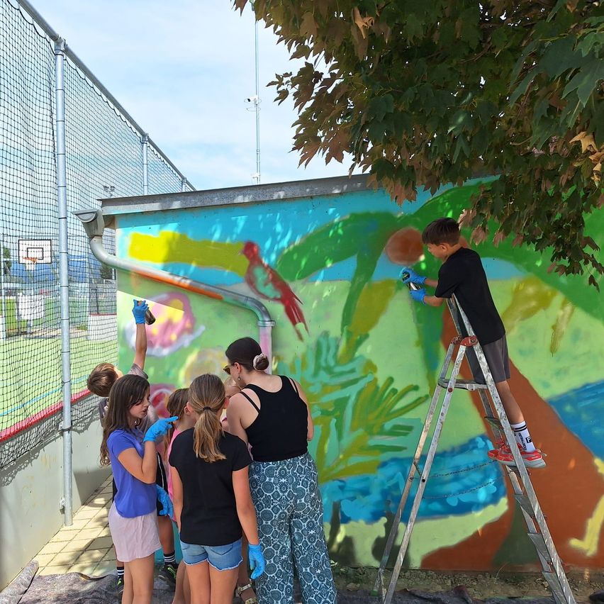 Eine Gruppe von Kindern und Erwachsenen malt ein farbenfrohes Wandbild. Das Bild zeigt eine tropische Szene mit Vögeln und Pflanzen. Sie benutzen Sprühfarbe und stehen auf Leitern und um die Wand herum.