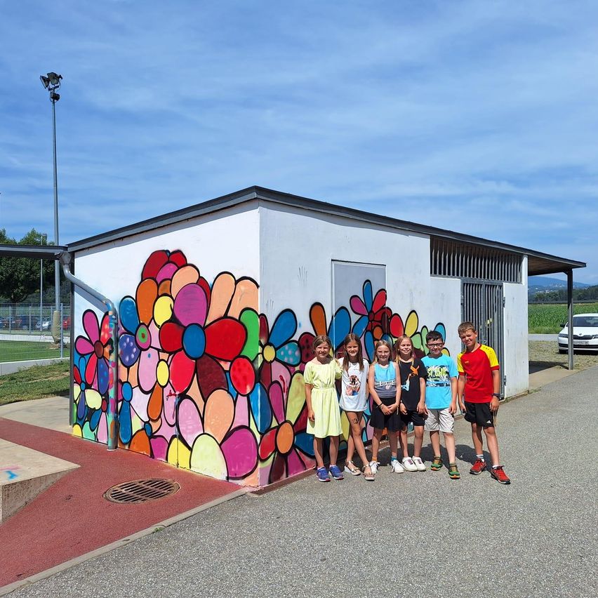 Kinder stehen vor einem Gebäude mit einem farbenfrohen Blumen-Mural an einem sonnigen Tag. Das Gebäude hat eine weiße Außenseite mit einem schwarzen Dach. Ein Straßenlicht steht hinter den Kindern.