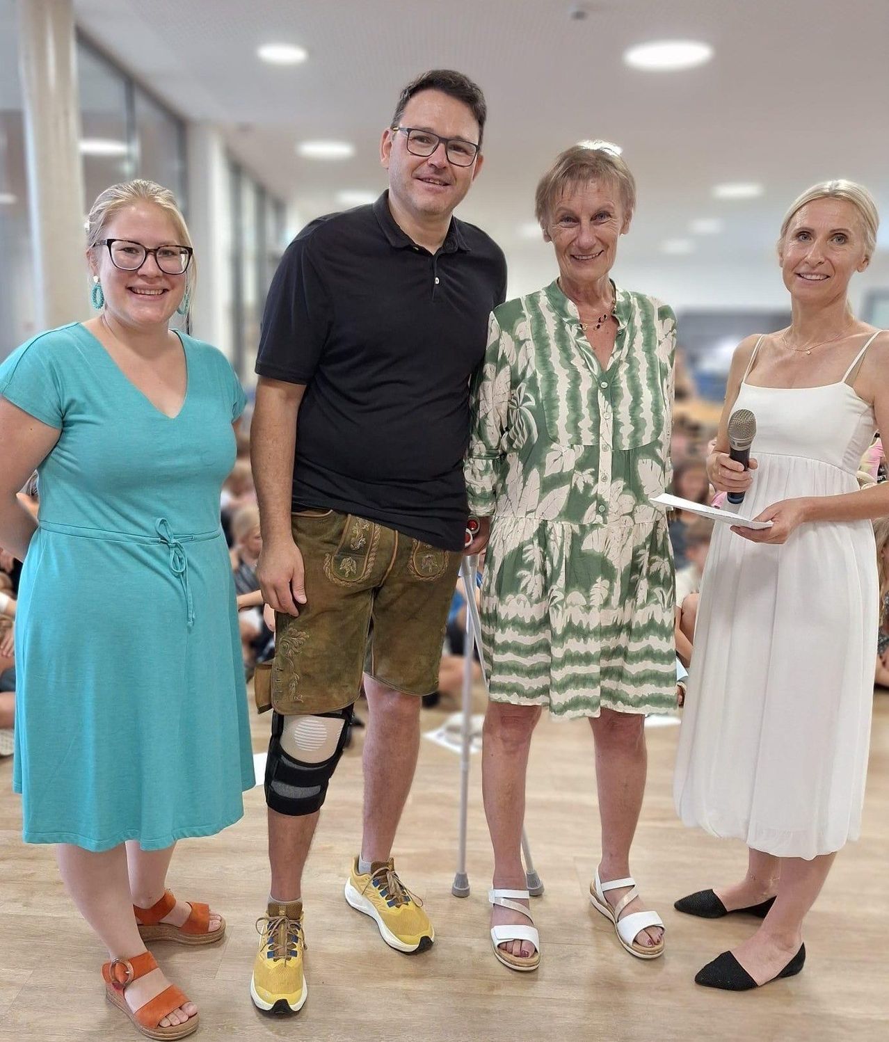 Vier Personen stehen lächelnd zusammen. Eine Frau hält ein Mikrofon, eine andere hat einen Stock und ein Mann trägt eine Kniebandage. Sie sind drinnen, wahrscheinlich bei einer Veranstaltung.