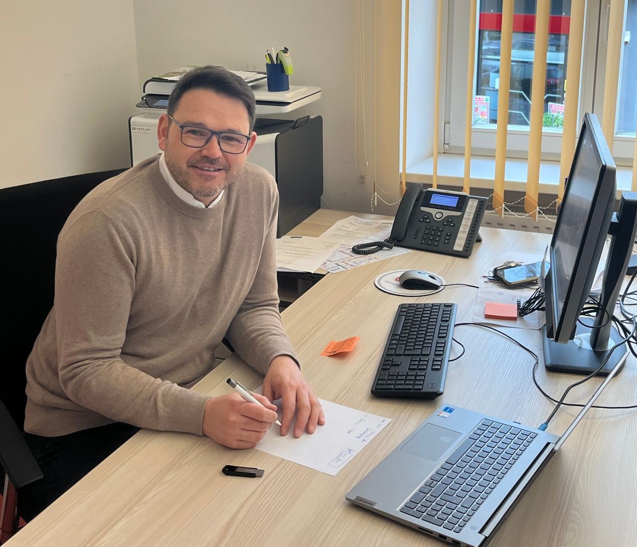 Ein Mann mit Brille und Pullover sitzt an einem Schreibtisch und schreibt auf Papier. Ein Laptop, eine Tastatur, eine Maus und ein Monitor sind auf dem Schreibtisch. Hinter ihm sind ein Drucker und ein Telefon zu sehen.