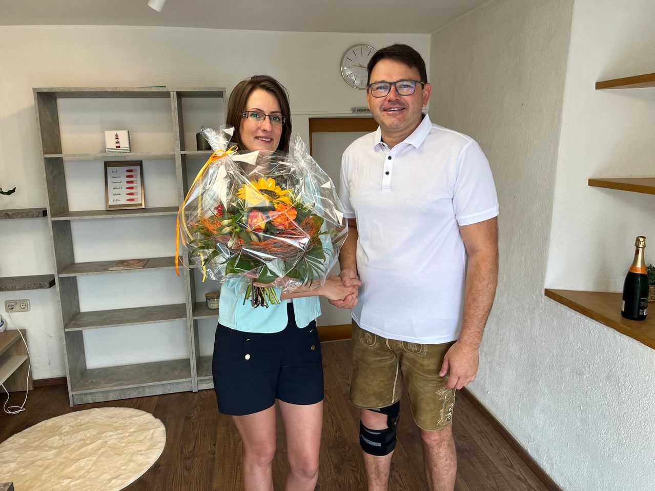 Ein Mann und eine Frau stehen in einem Raum und halten Händchen. Die Frau hält einen Blumenstrauß. Hinter ihnen steht ein Regal mit verschiedenen Gegenständen. Eine Uhr ist an der Wand montiert.