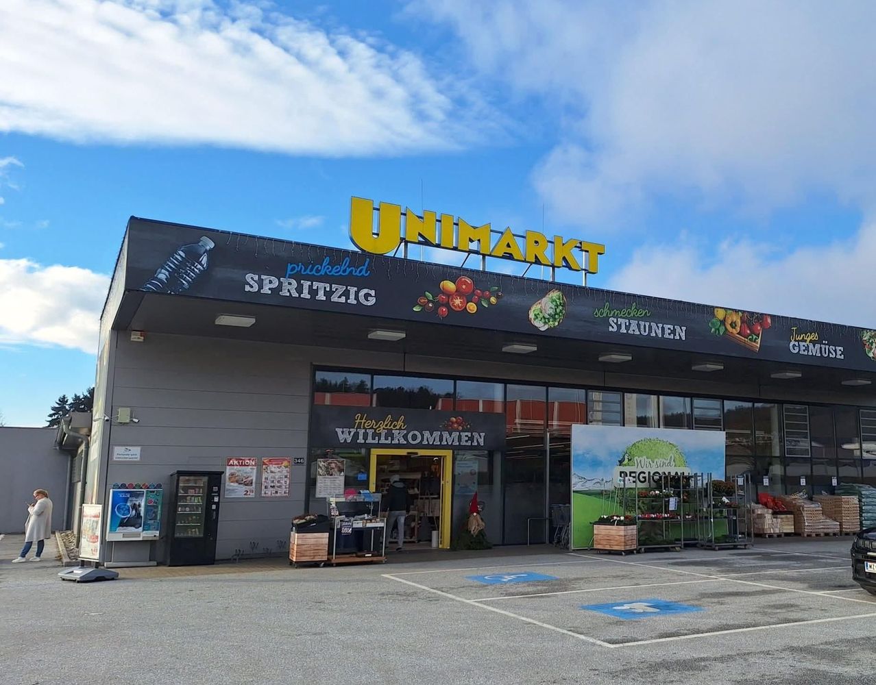 Außenansicht eines Unimarkt-Geschäfts mit blauem Himmel und Wolken. Ein gelbes Schild zeigt Unimarkt. Blumen sind draußen.