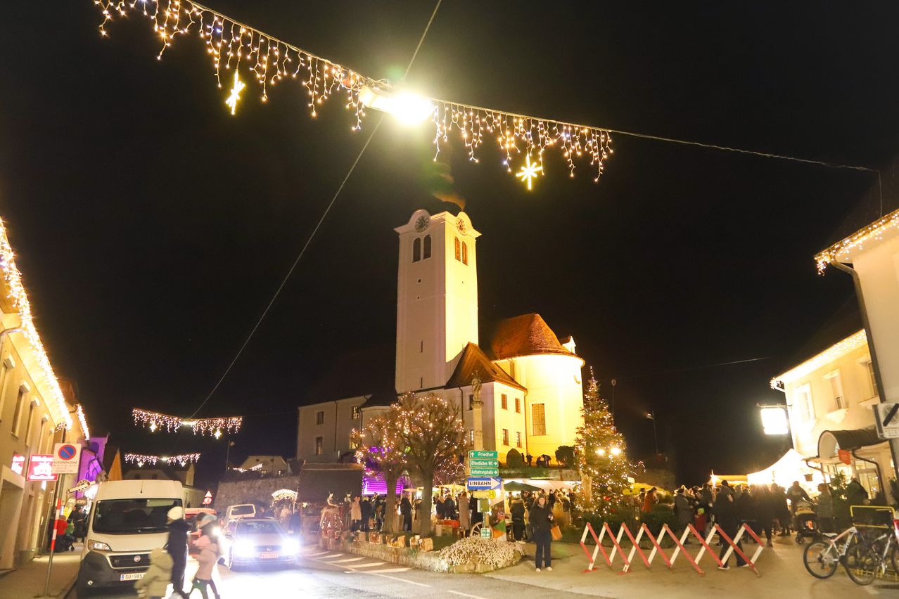 Eine Menschenmenge versammelt sich bei Nacht vor einer Kirche während der Feiertage. Die Kirche ist mit Weihnachtslichtern und Bäumen geschmückt.