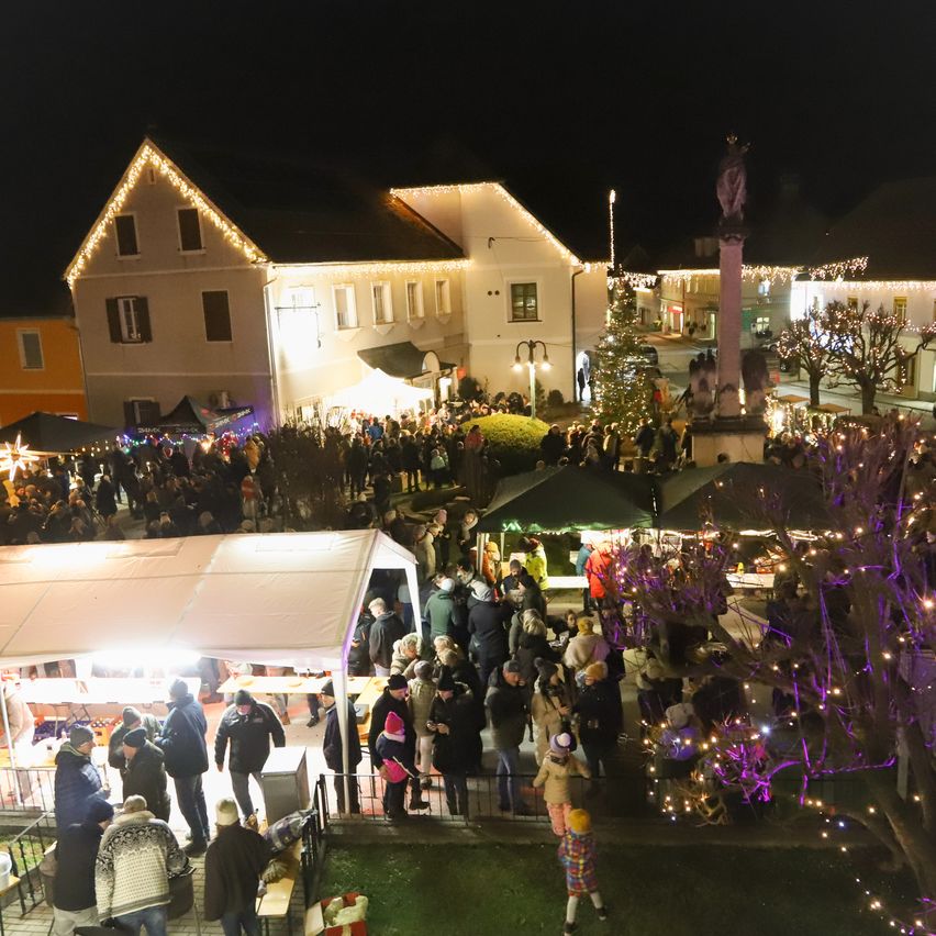 Eine große Menschenmenge versammelt sich nachts in einem Freiluftbereich, geschmückt mit Lichtern und Weihnachtsdekoration. Menschen gehen umher, einige in Winterkleidung. Es gibt mehrere Stände mit Zelten und einem Weihnachtsbaum im Hintergrund.