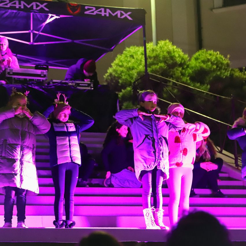 Kinder, die Winterkleidung tragen und Rehenhörner auf dem Kopf haben, stehen auf einer Bühne mit lila Lichtern. Dahinter sitzt ein DJ unter einem Zelt. Ein kleines Publikum sitzt auf den Stufen.