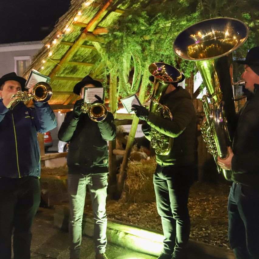Vier Musiker spielen ihre Blechblasinstrumente vor einer Hütte mit einem schneebedeckten Dach. Sie tragen Winterkleidung und Mützen.