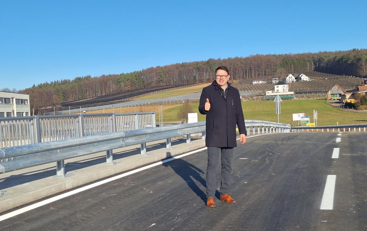 Ein Mann steht auf einer Straße, gibt ein Daumen hoch und hat im Hintergrund einen Zaun und Bäume.