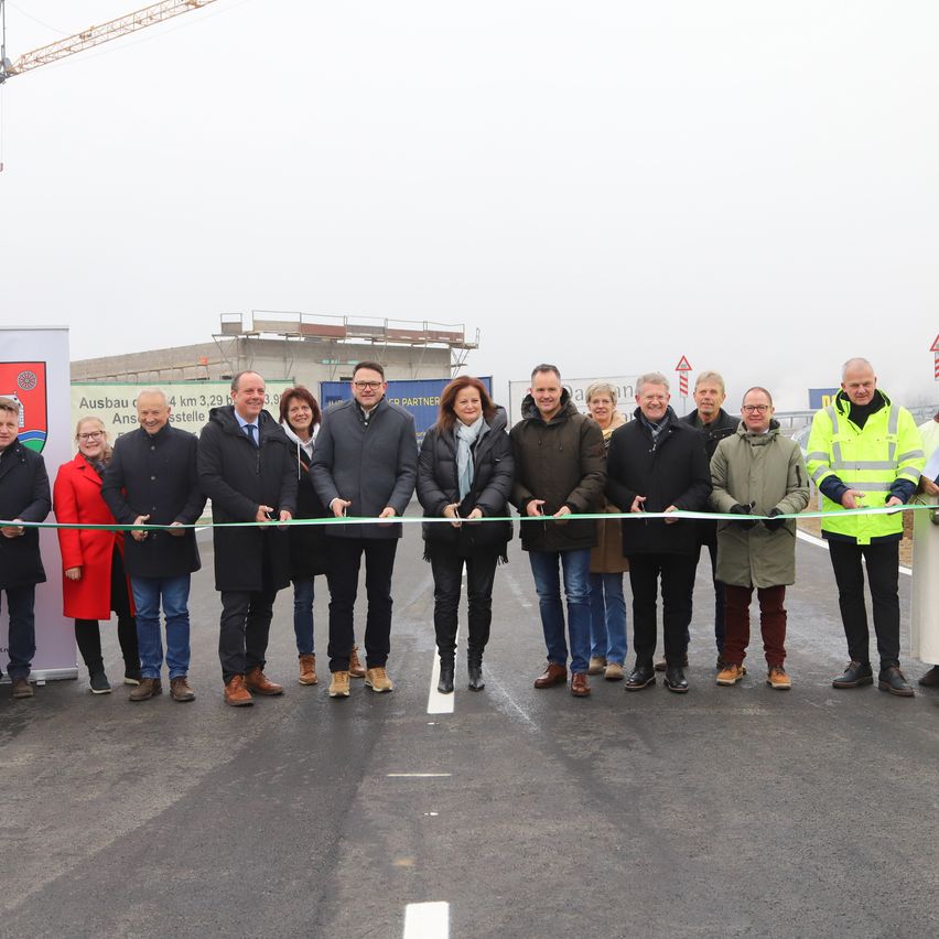 Eine Gruppe von Menschen in formeller Kleidung steht in einer Reihe, hält ein Band und feiert eine Eröffnungsveranstaltung. Dahinter ist ein Bau zu sehen.